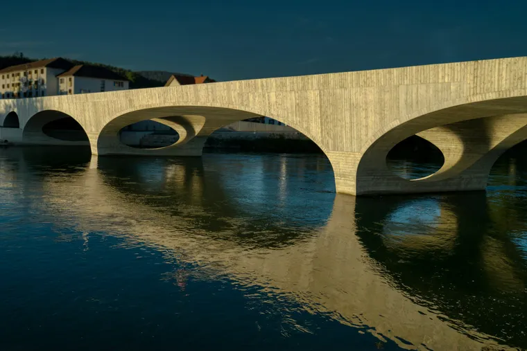 Betonbogenbrücke über die Aare in Aarau im Abendlicht, die sich im Wasser spiegelt, dahinter Häuser am Ufer
