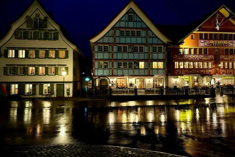 Bemalte Häuser im Dorfkern von Appenzell am Abend, Lichter spiegeln sich auf dem nassen Pflaster